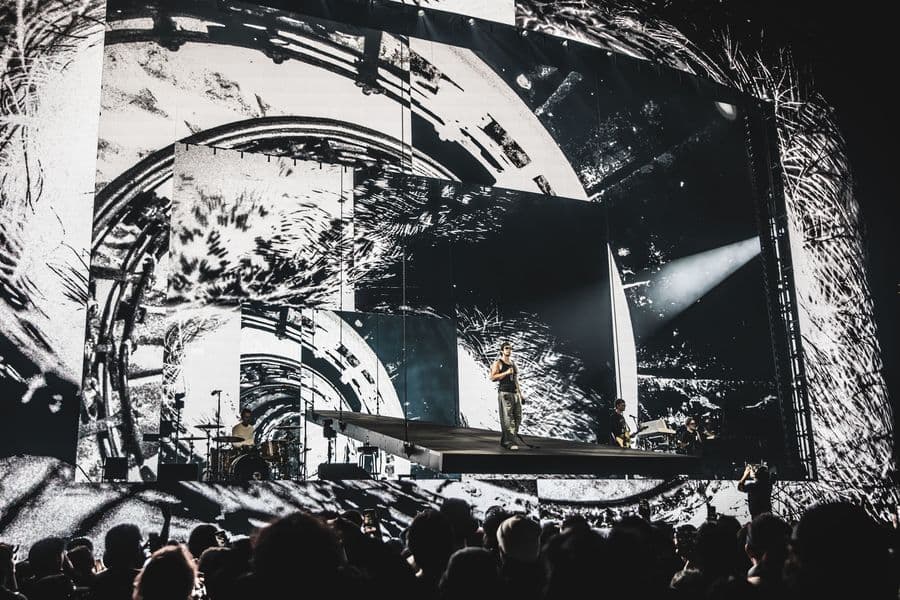 Joji standing on a suspended stage runway that extends out toward the audience. The massive LED backdrop displays dynamic, custom video content designed by Fray Studio, featuring a high-contrast, black-and-white gritty visual of distorted mechanical gears and swirling, abstract textures. His live band plays on the main stage behind him, with a silhouetted crowd watching from the foreground.
