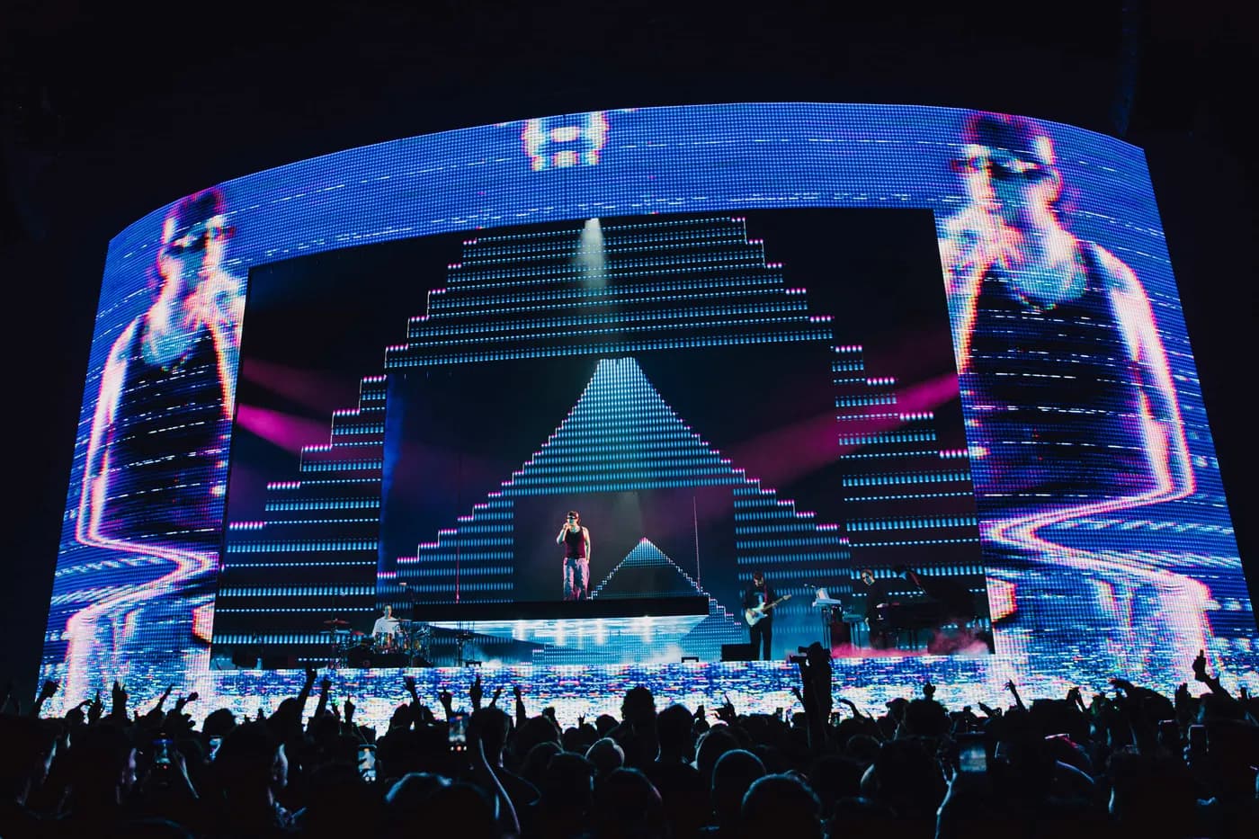 Joji performing on a raised central stage platform in front of a massive curved LED screen. The screen displays dynamic video content designed by Fray Studio, featuring a digital, glitch-art style blue and pink stepped pyramid flanked by two giant, distorted projections of Joji. A live band plays in the background, and a silhouetted crowd cheers in the foreground.