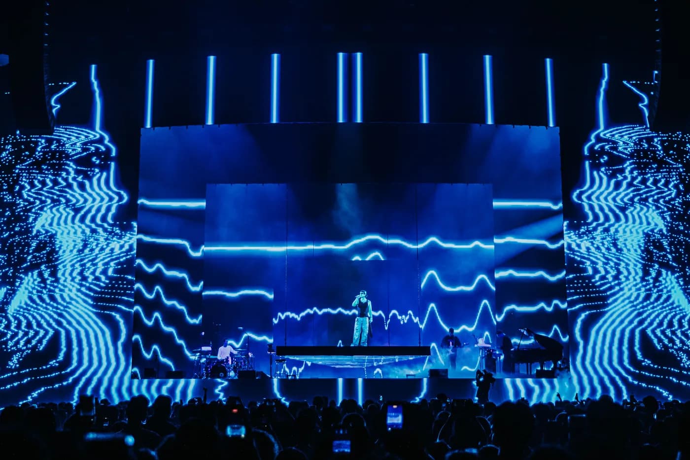 Joji singing center stage accompanied by his live band, surrounded by giant screens displaying vibrant, electric blue, oscillating horizontal lines. This luminous, soundwave-style video content was designed by Fray Studio. A silhouetted crowd is visible holding up phones in the foreground.