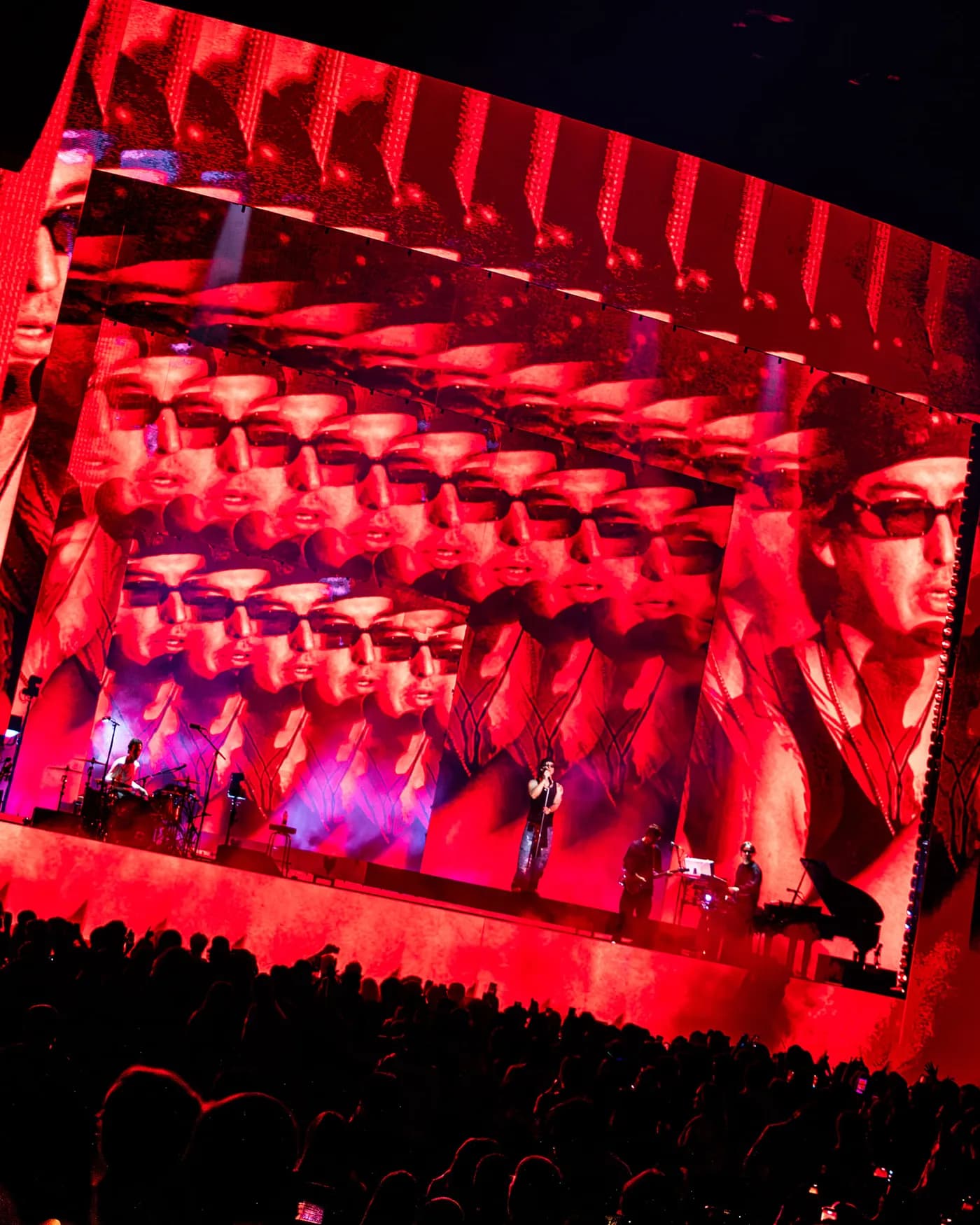 Joji performing on stage with his band, illuminated by intense red lighting. The massive backdrop features custom video content by Fray Studio, displaying a distorted, repeating grid of Joji's face wearing sunglasses. A large crowd is visible in silhouette in the foreground.