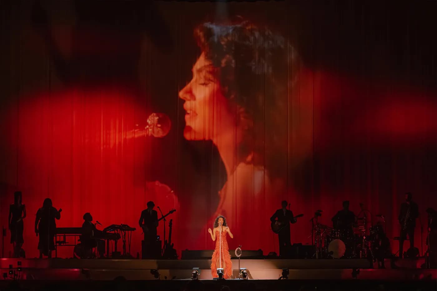 Olivia Dean performing center stage in a shimmering red-orange dress with a feathered hem, illuminated by a bright spotlight. Behind her and her silhouetted live band, a massive backdrop curtain displays custom video projection designed by Fray Studio. The cinematic visuals feature a giant, vintage-style profile of a woman singing into a microphone, bathed in deep, atmospheric red light.