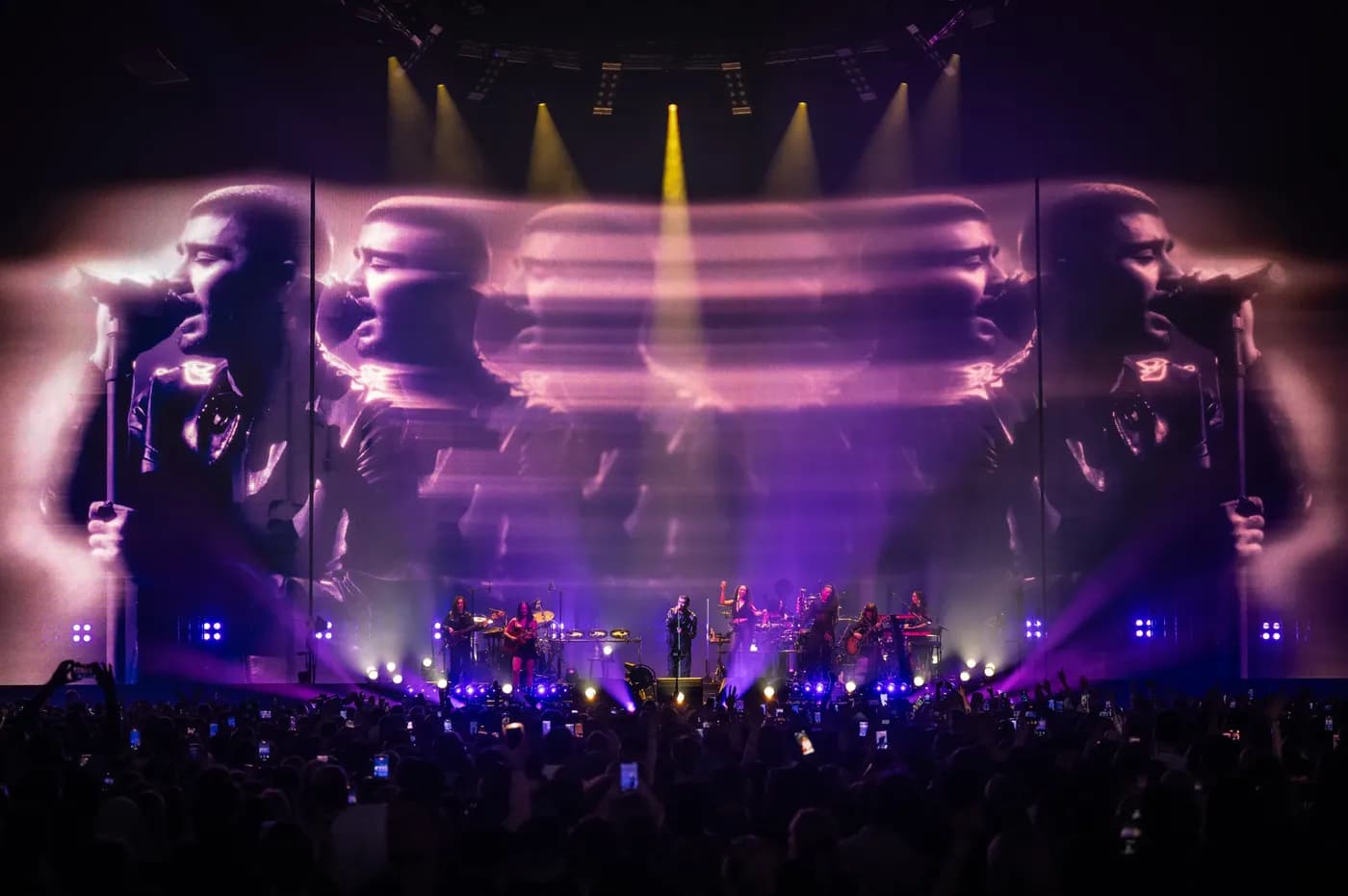 Zayn Malik performs on stage with his band in front of a massive digital screen displaying a repeating, purple-streaked panorama of his performance.