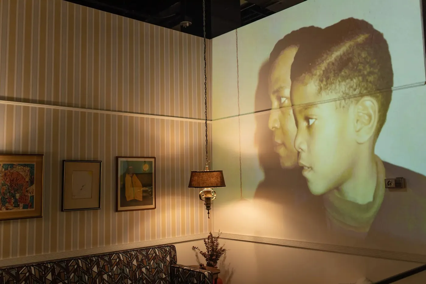 Projected photograph of Basquiat and child on striped wall at King Pleasure exhibition, with framed art and lamp in foreground.