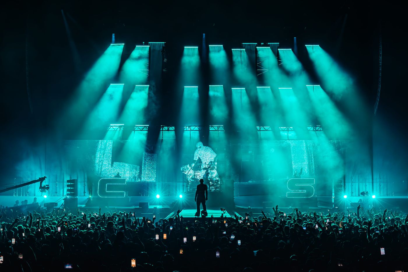 Chase and Status 2025 tour live: silhouette of MC on catwalk before cheering O2 London crowd, green‑teal beams spotlight graffiti‑style backdrop.