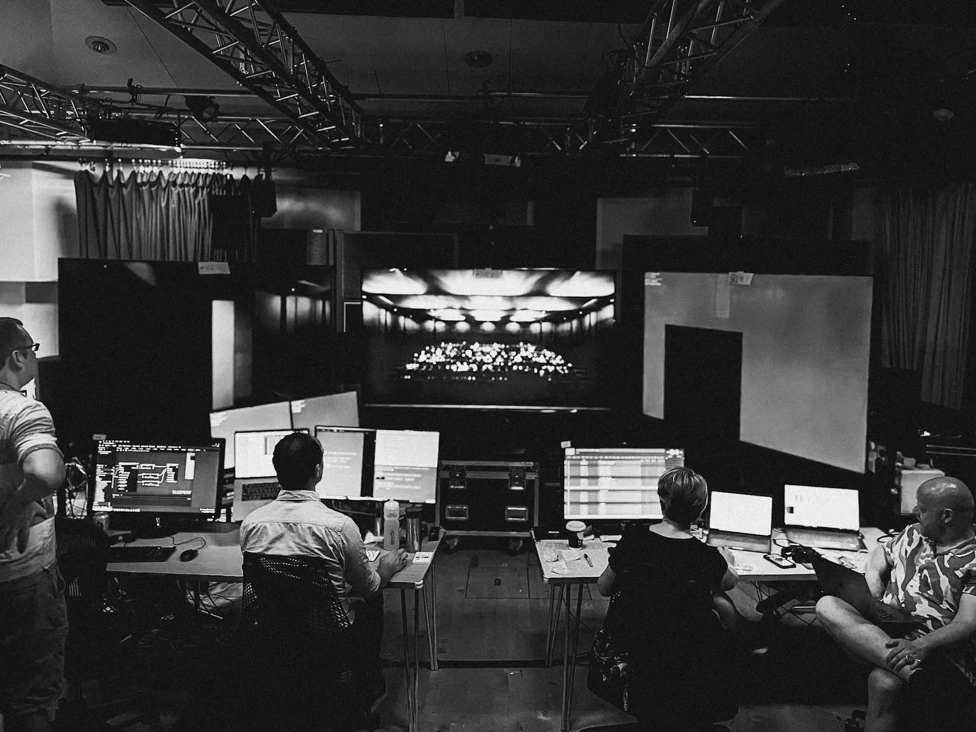 4 people gathered around computers discussing an idea with a large screen in the back ground