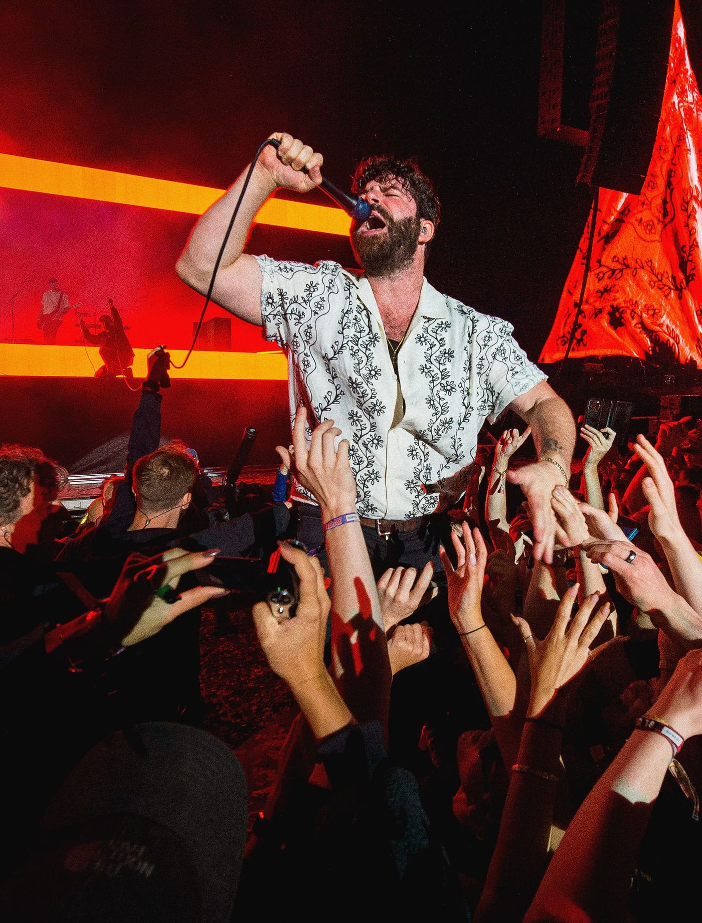 Foals singer Yannis Philippakis leans into the crowd mid-song, with FRAY Studio’s red stage visuals behind him.
